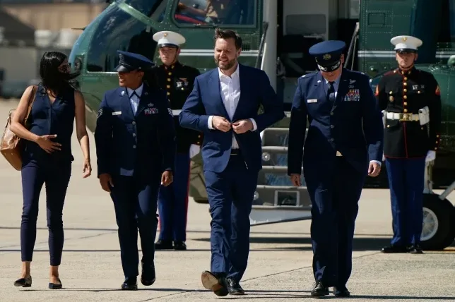 Muškarac u plavom odijelu popravlja zakopčano dugme dok hodaju sa ženom u tamnoplavoj odjeći i dve osobe u vojnim uniformama, na aerodromu pored helikoptera.