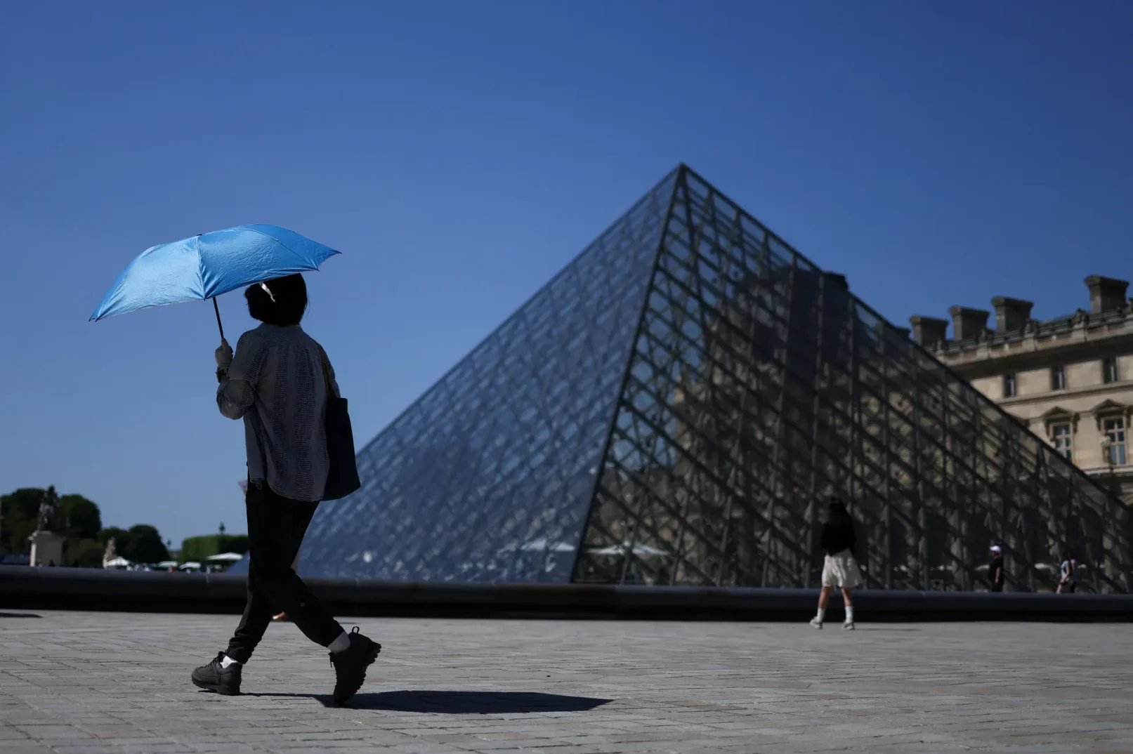 Osoba s plavim kišobranom šeta ispred Piramide Louvre u Parizu.