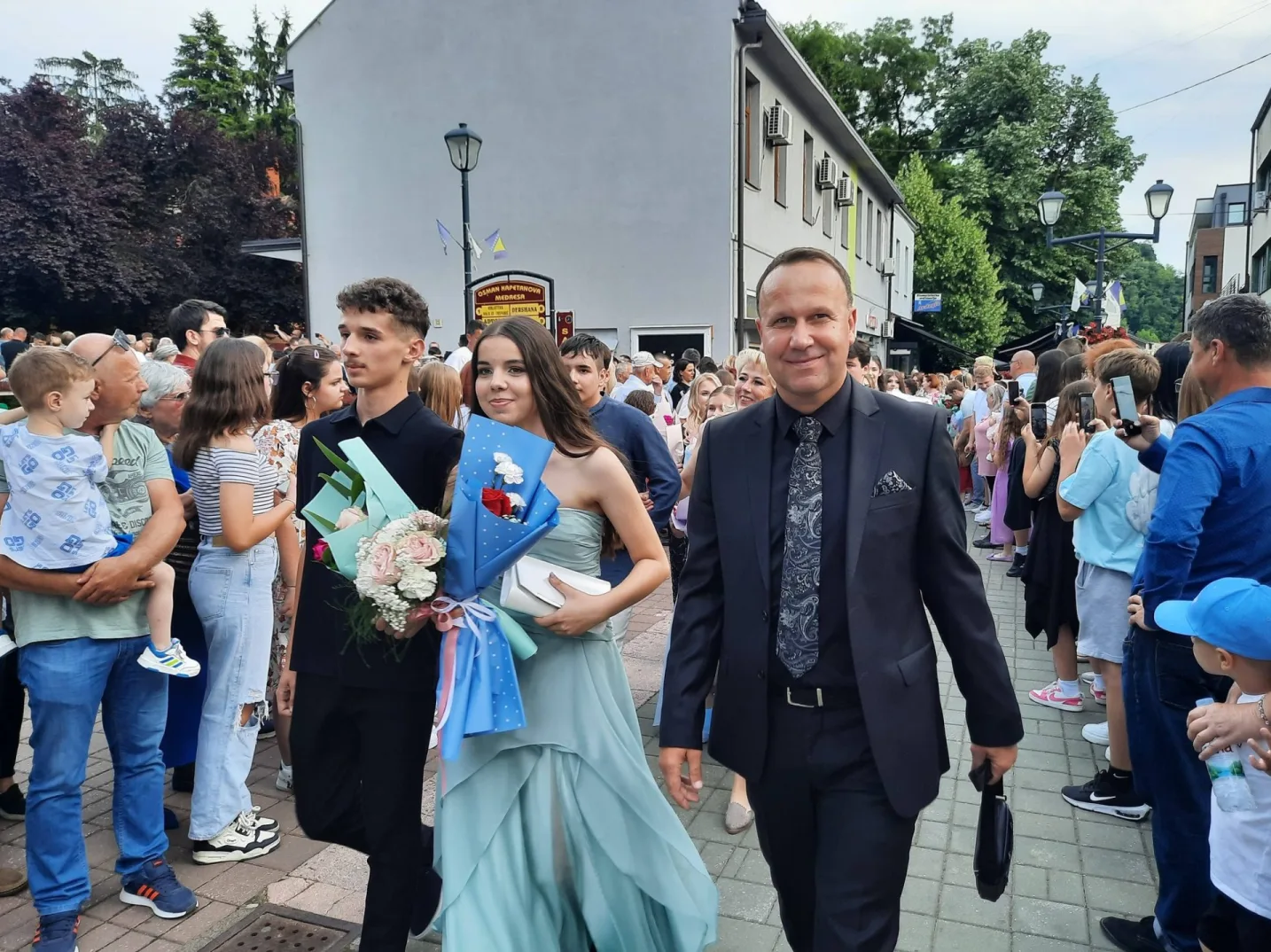 Maturanti i njihovi roditelji slave matursku veče na ulici, nose cvijeće i uživaju u svečanom trenutku.