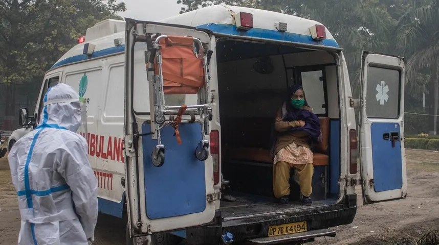 Medicinska sestra u zaštitnom odijelu stoji ispred otvorenih zadnjih vrata ambulantnih kola, dok pacijentica sjedi unutra noseći masku za lice.