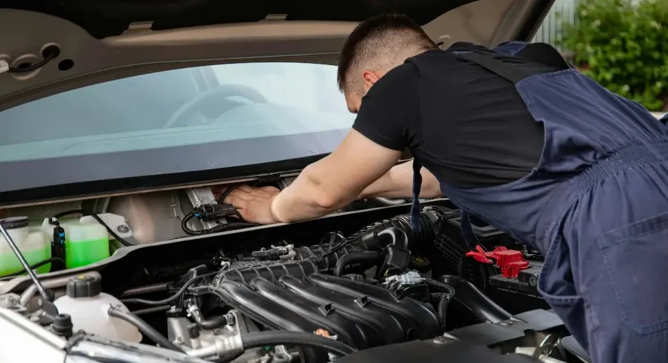Mehanicar u plavom kombinezonu radi na motoru automobila, pregleda ili popravlja dijelove ispod haube.
