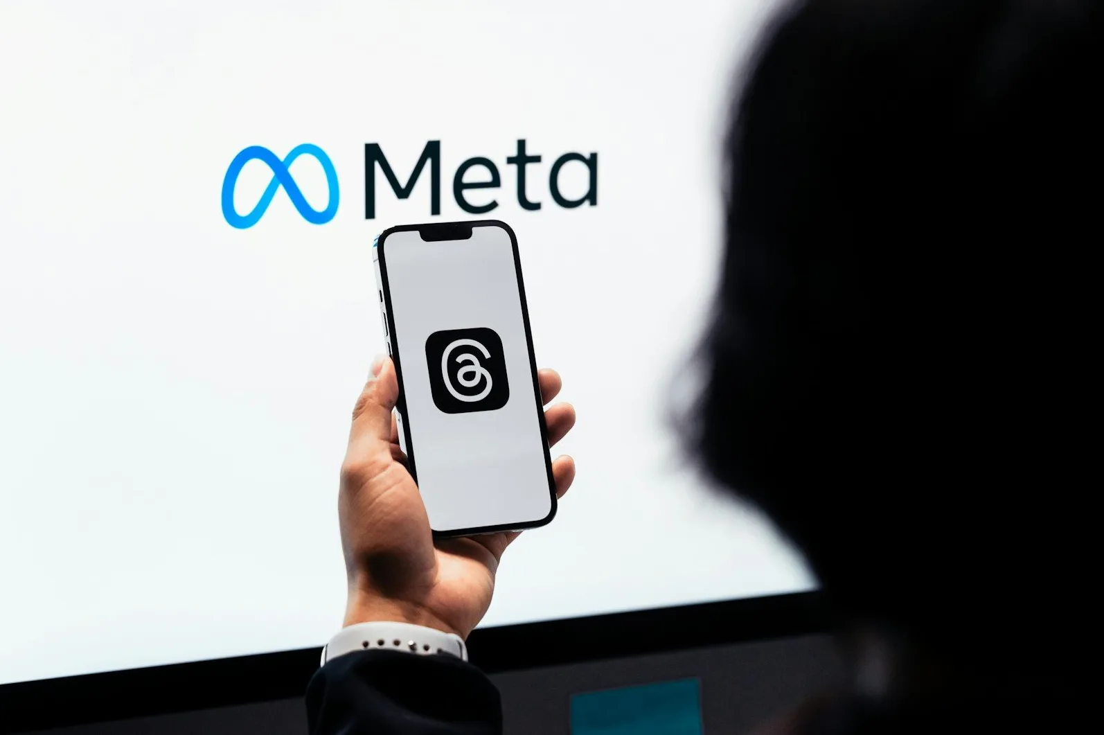 Osoba drži pametni telefon s prikazanom aplikacijom "Threads" ispred velikog ekrana sa logotipom "Meta" i natpisom.