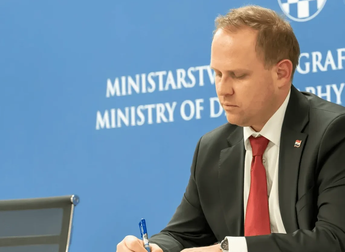 Ministar u crnom odijelu i crvenoj kravati piše olovkom, s pozadinom na kojoj piše "MINISTRY OF DEFENCE" i hrvatskim grbom.