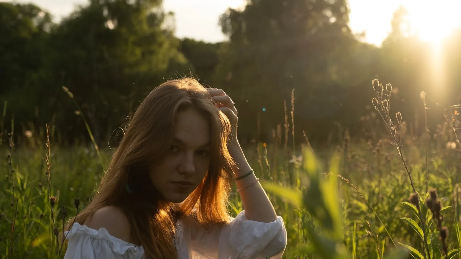 Portret mlade djevojke sa dugom smeđom kosom, obučena u bijelu haljinu otvorenih ramena, kako sjedi u polju trave i cvijeća tokom zalaska sunca. Sunce probija kroz drveće u pozadini.