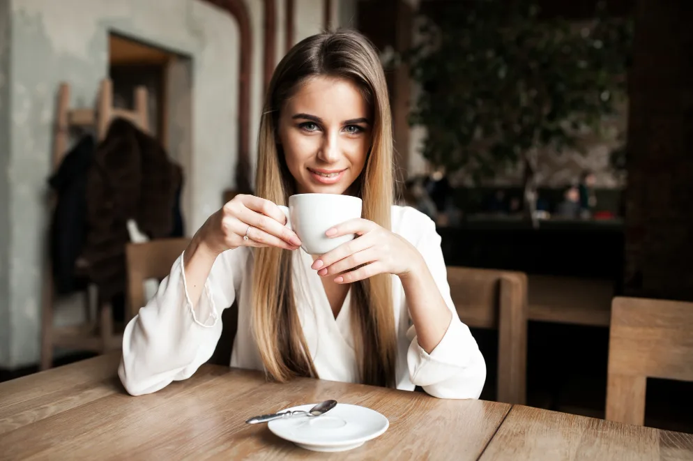 Mlada žena u bijeloj košulji drži bijelu šoljicu kafe i smiješi se u kameru, sjedeći za drvenim stolom u kafiću.