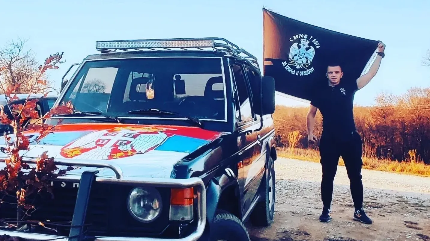 Mladić u crnoj majici i pantalonama drži crnu zastavu sa grbom dvoglavog orla i natpisom na ćirilici, stojeći pored terenskog vozila koje je ukrašeno srpskom trobojkom.