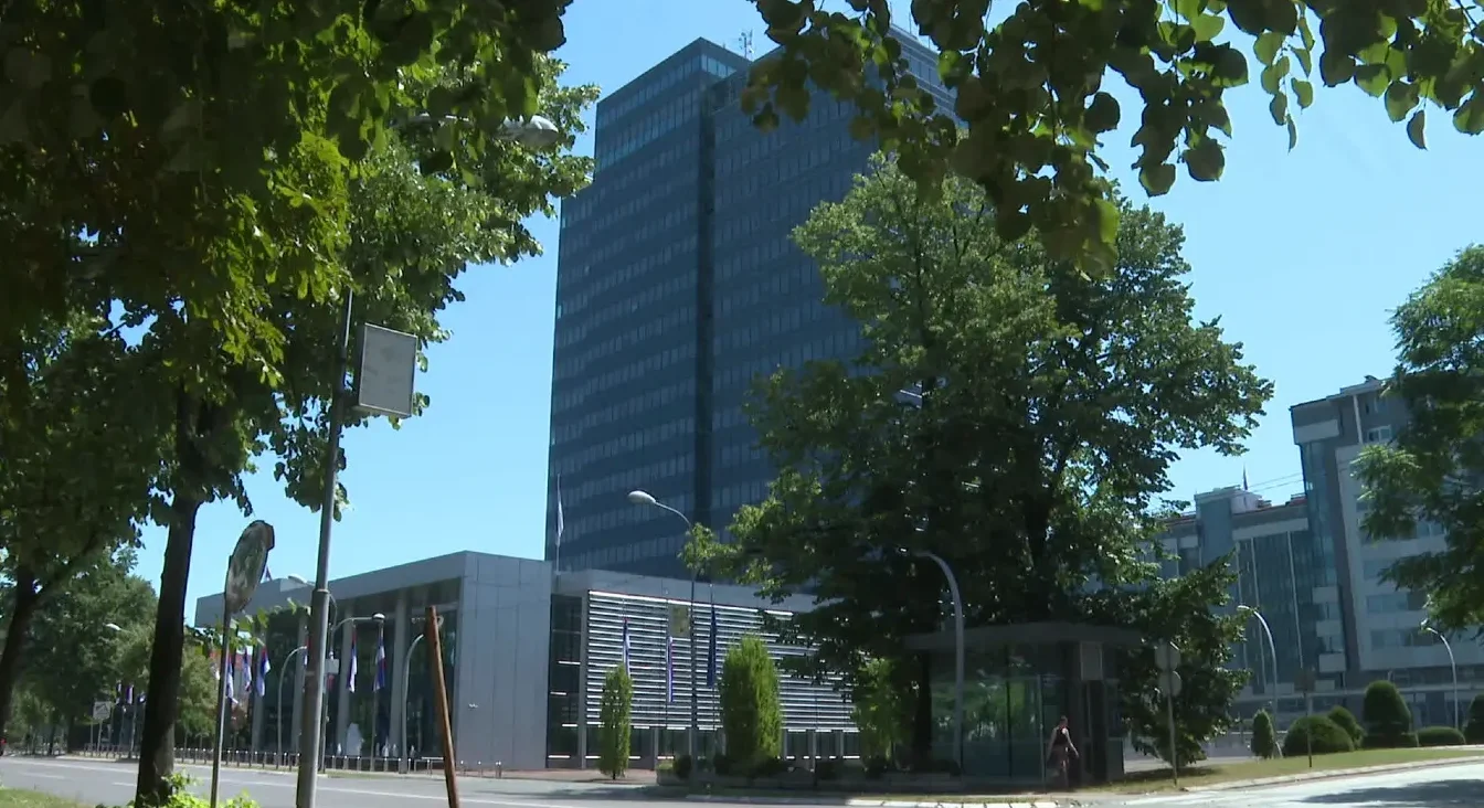 Moderan gradjevinski kompleks sa visokom staklenom zgradom u centru i drvećem ispred.