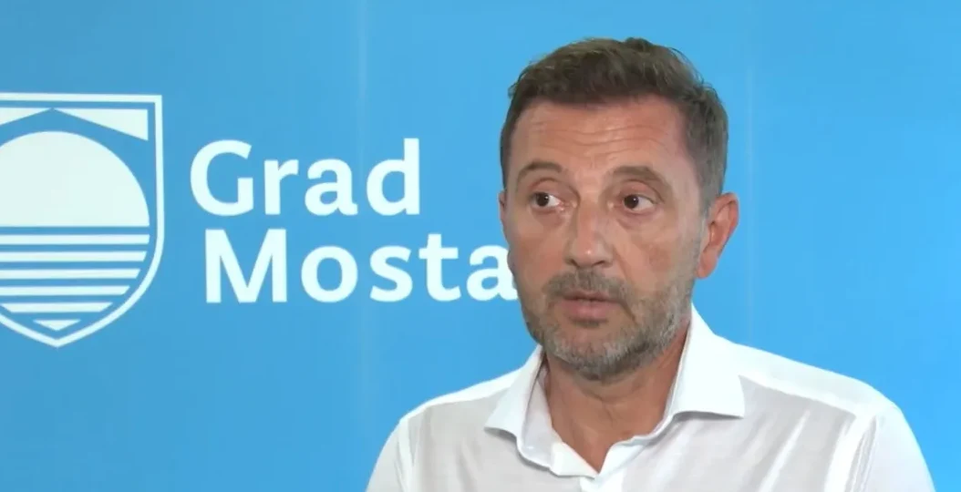 Muškarac u bijeloj košulji ispred plave pozadine sa grbom i natpisom Grad Mostar