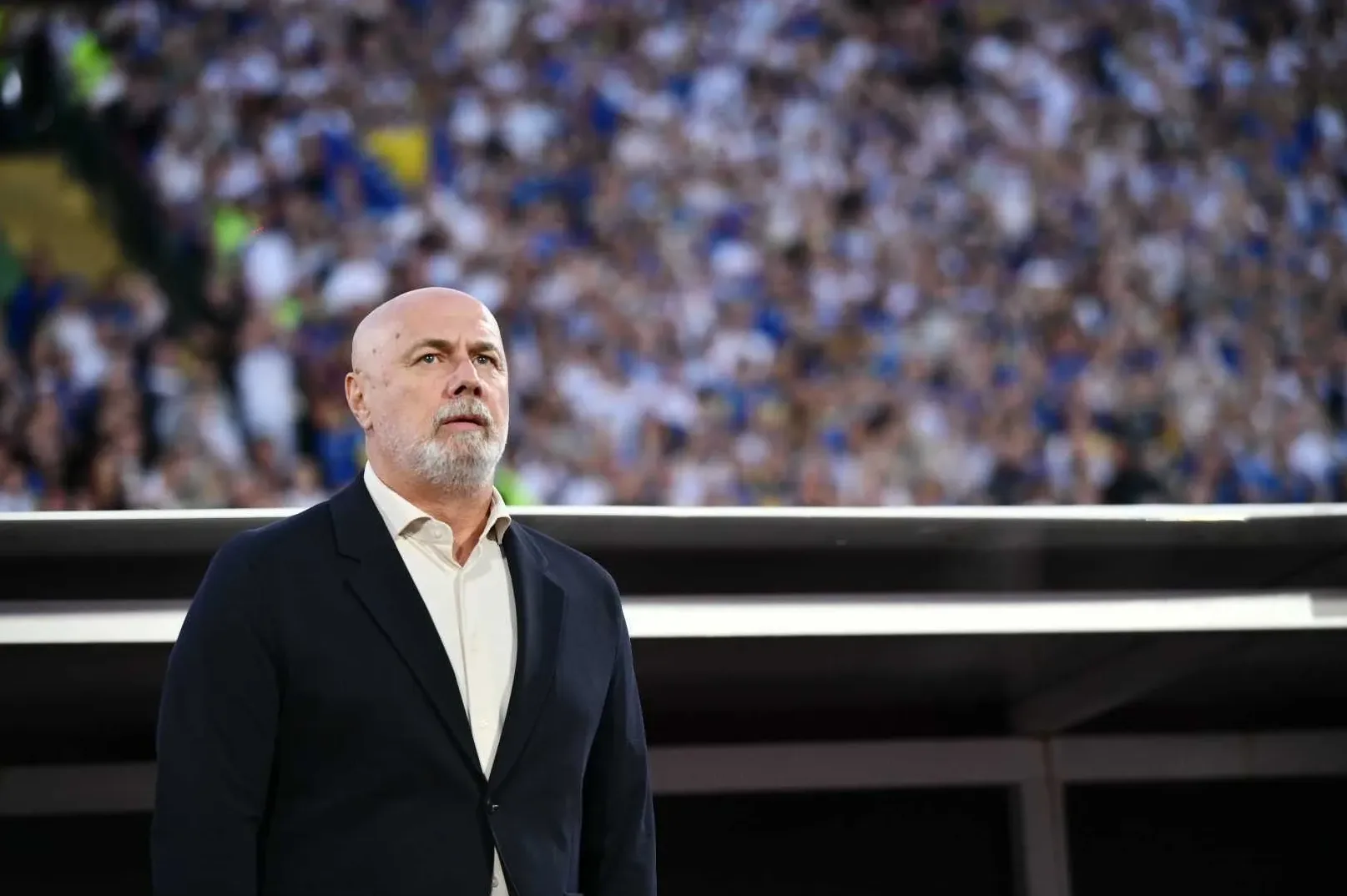 Ćelava glava muškarca sa sijedom bradom u tamnom sakou gleda u daljinu, sa zamagljenim tribinama prepunim navijača u pozadini.