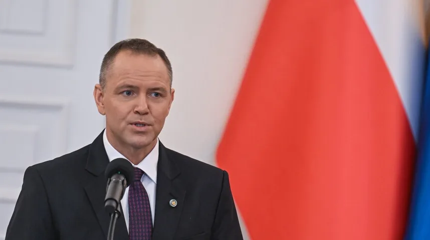 Muškarac u odijelu govori iza mikrofona, s poljskom zastavom u pozadini.