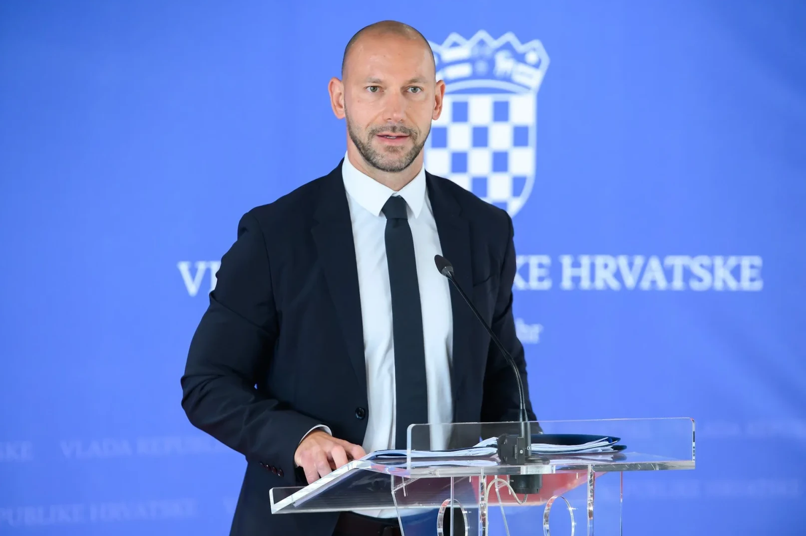 Muškarac u tamnom odijelu i bijeloj košulji sa crnom kravatom stoji iza prozirnog podijuma sa mikrofonom.