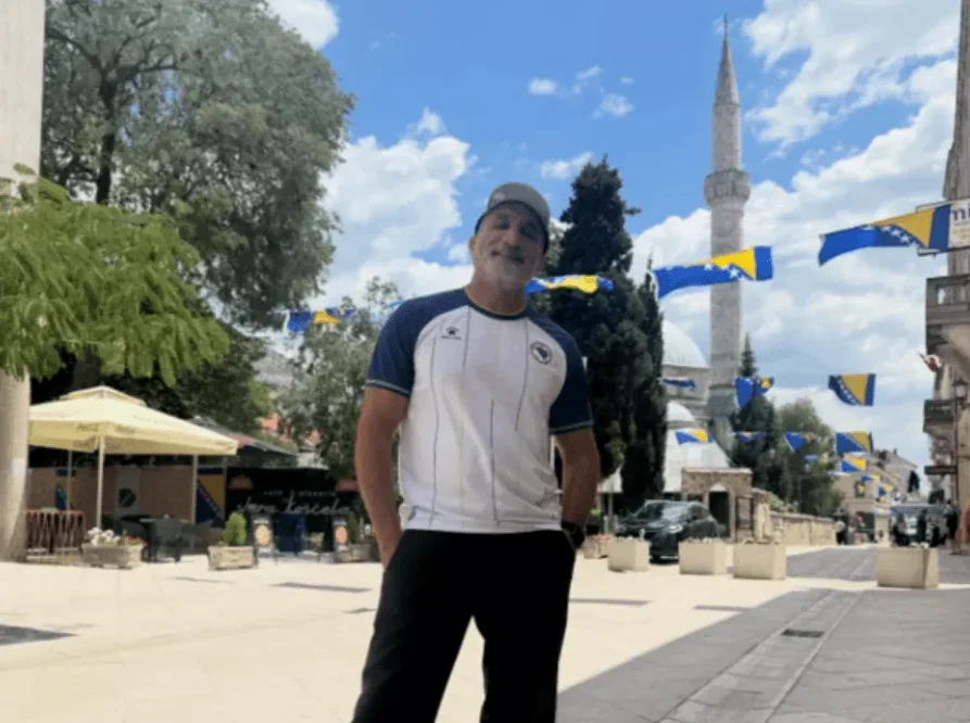 Muškarac u bijeloj majici sa plavim rukavima i šiltericom pozira na sunčanom trgu ukrašenom zastavama Bosne i Hercegovine, s džamijom u pozadini.