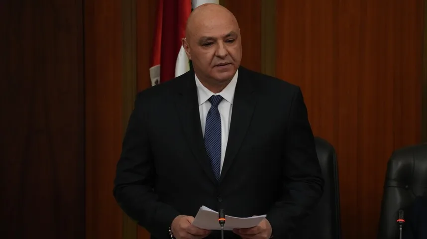 Muškarac u crnom odijelu i plavoj kravati drži papire i govori ispred mikrofona u instituciji.