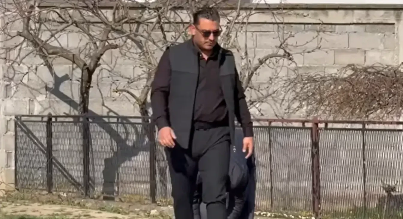 Mlađi muškarac obučen u crnu košulju, prsluk, pantalone i sunčane naočale, sa torbom preko ramena, hoda uz ogradu i drveće bez lišća.
