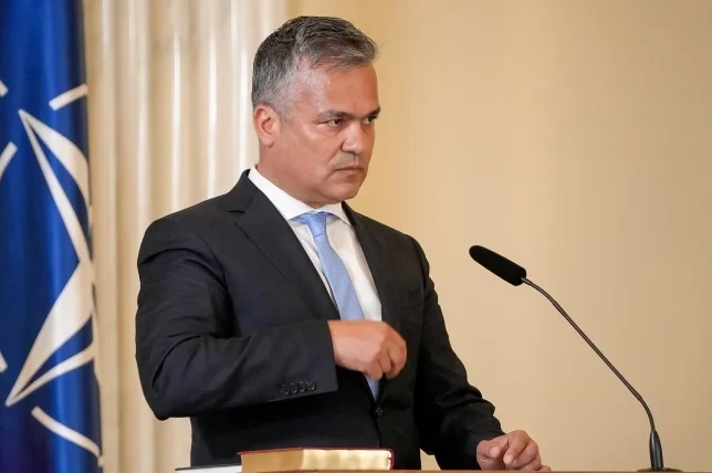 Političar u tamnom odijelu, bijeloj košulji i plavoj kravati, drži govor iza drvene govornice pored zastave NATO-a.