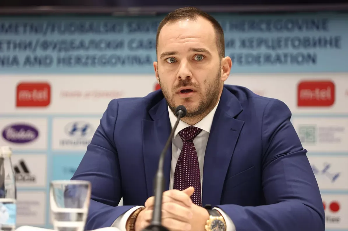 Muškarac u plavom odijelu i ljubičastoj kravati govori na konferenciji za medije, s logotipom Nogometnog/Fudbalskog saveza Bosne i Hercegovine u pozadini.