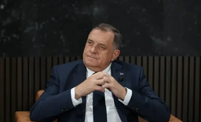 Političar u tamnom odijelu i bijeloj košulji, sa tamnom kravatom, sjedi i gleda lijevo.