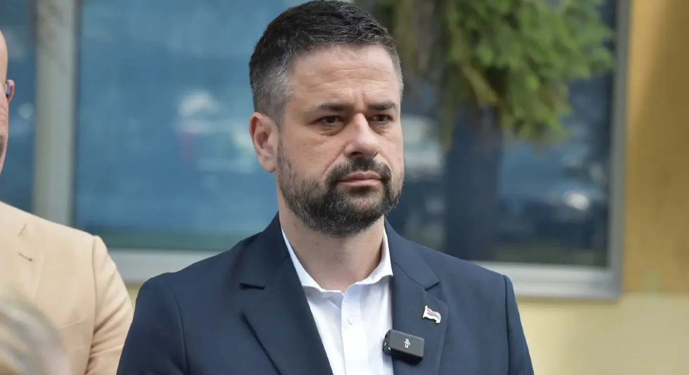 Muškarac u tamnom odijelu i bijeloj košulji, sa bratskim amblemom na reveru, gleda u kameru.