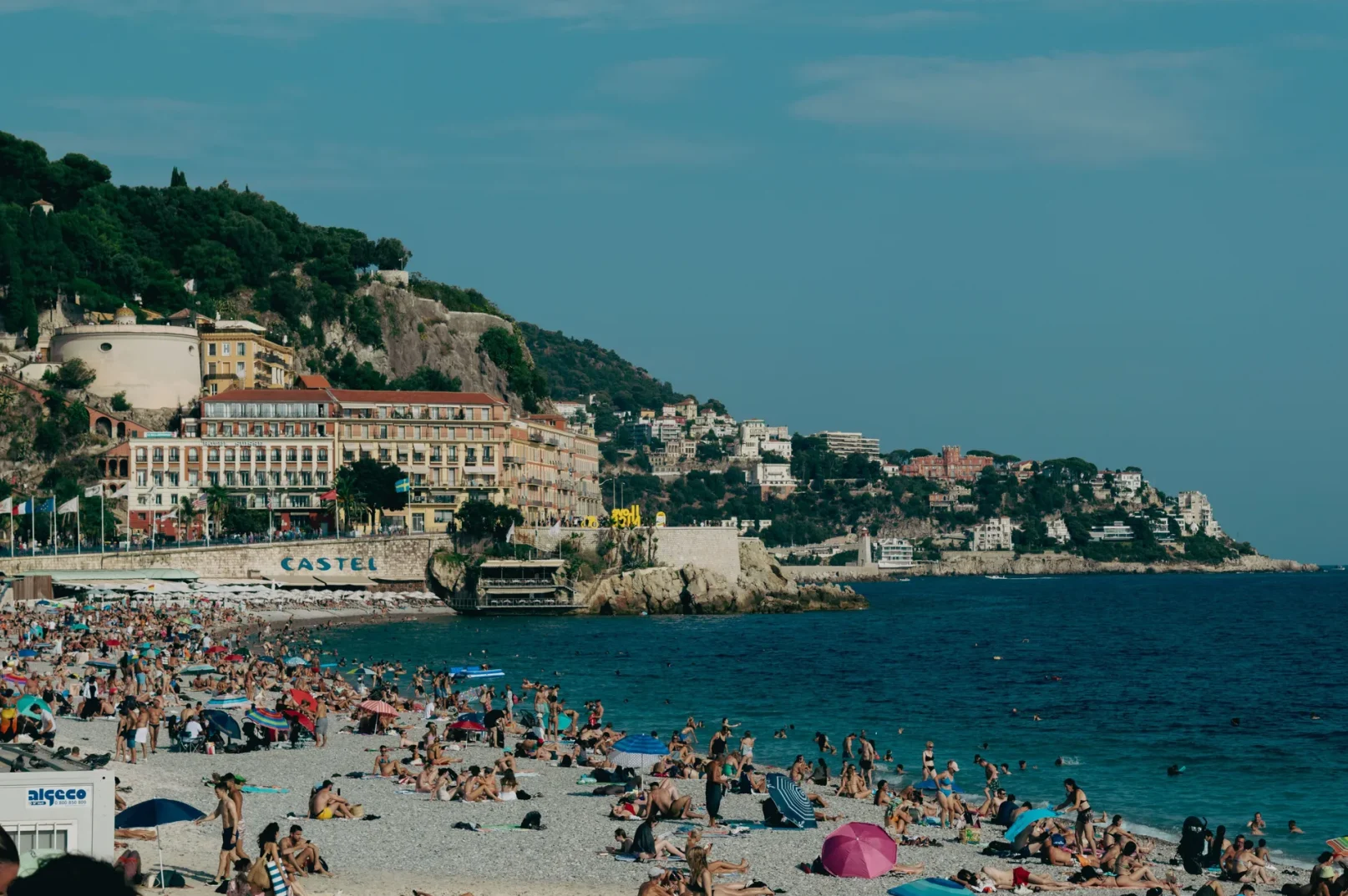 Prepun kameniti pejzaž plaže sa mnogo ljudi koji se sunčaju i kupaju u plavom moru, sa zgradama i zelenilom na brdu u pozadini.