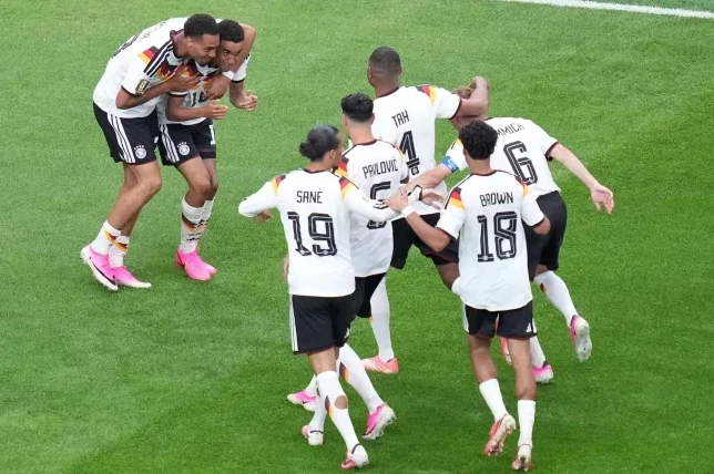 Fudbaleri Njemačke u bijelim dresovima sa crnim i zlatnim detaljima grle se i slave gol na zelenom fudbalskom terenu.