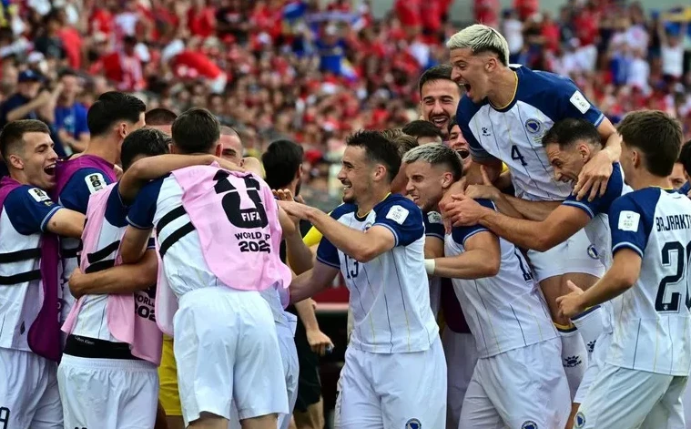 Fudbalska reprezentacija Bosne i Hercegovine u bijelim dresovima slavi na terenu, okruženi navijačima u pozadini.