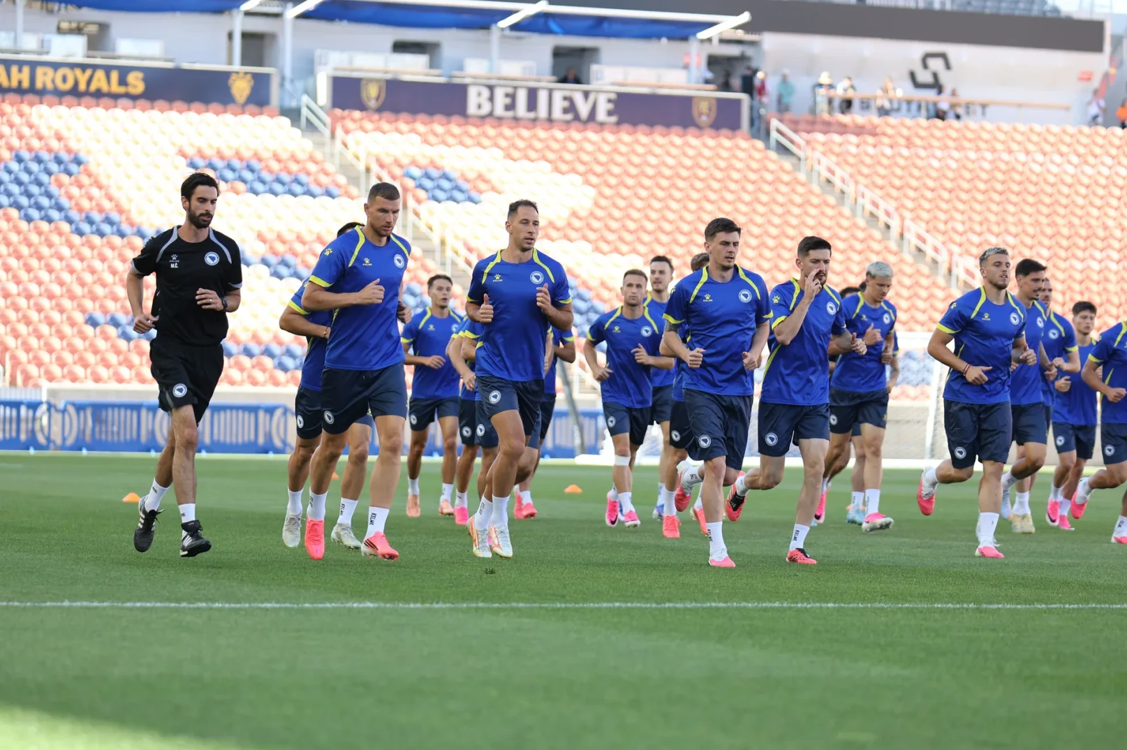 Fudbaleri u plavim dresovima trče na zelenom terenu stadiona, sa tribinama u pozadini.