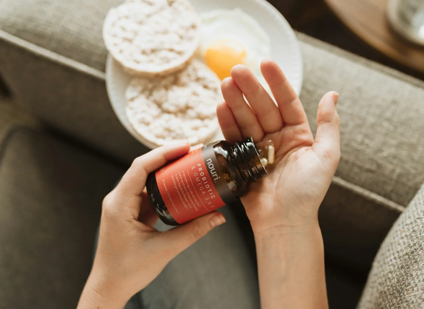 Žena drži bocu Nouri Probiotic & Omega 3,6,9 suplemenata, s nekoliko kapsula u ruci, pored tanjira s jajetom i rižinim galetama.