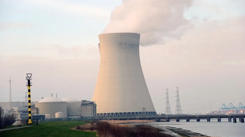 Veliki rashladni toranj nuklearne elektrane sa parom koja izlazi iz vrha, uz obalu sa stubovima za prenos električne energije i brodovima u luci u daljini.