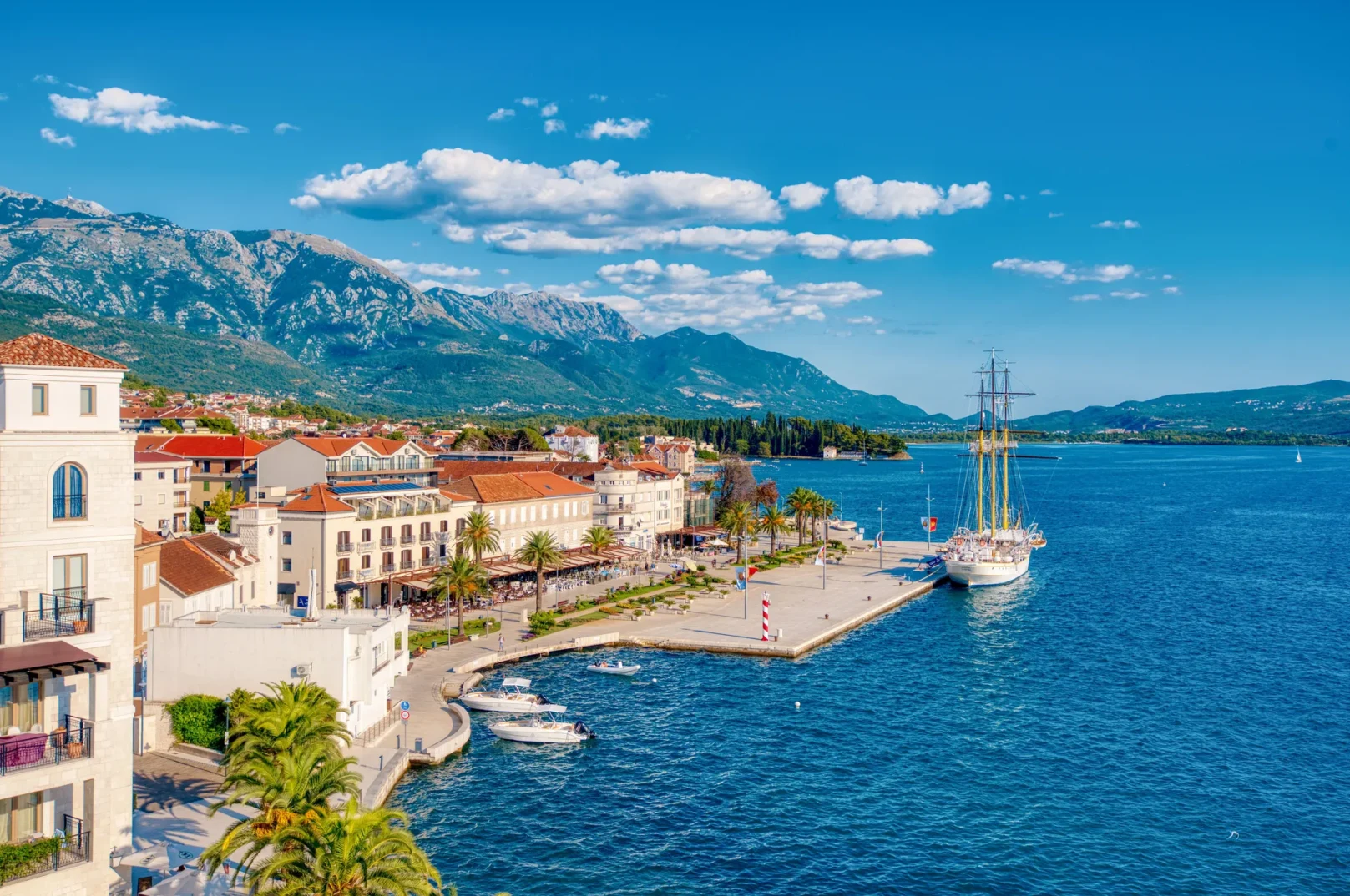 Pogled na mediteransko ljetovalište s bijelim zgradama, palmama i planinama u pozadini, kao i prekrasnim jedrenjakom usidrenim uz obalu mora.