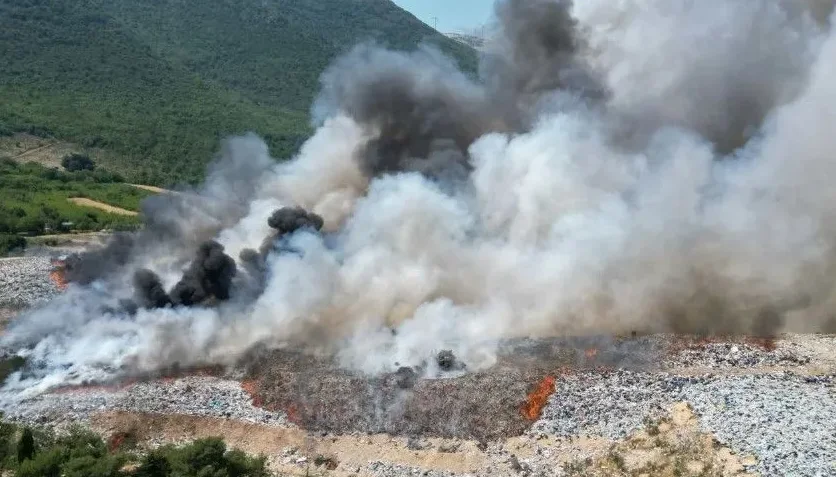 Velika deponija smeća u plamenu sa gustim oblacima crnog i bijelog dima koji se dižu u zrak, okružena zelenim brdima.