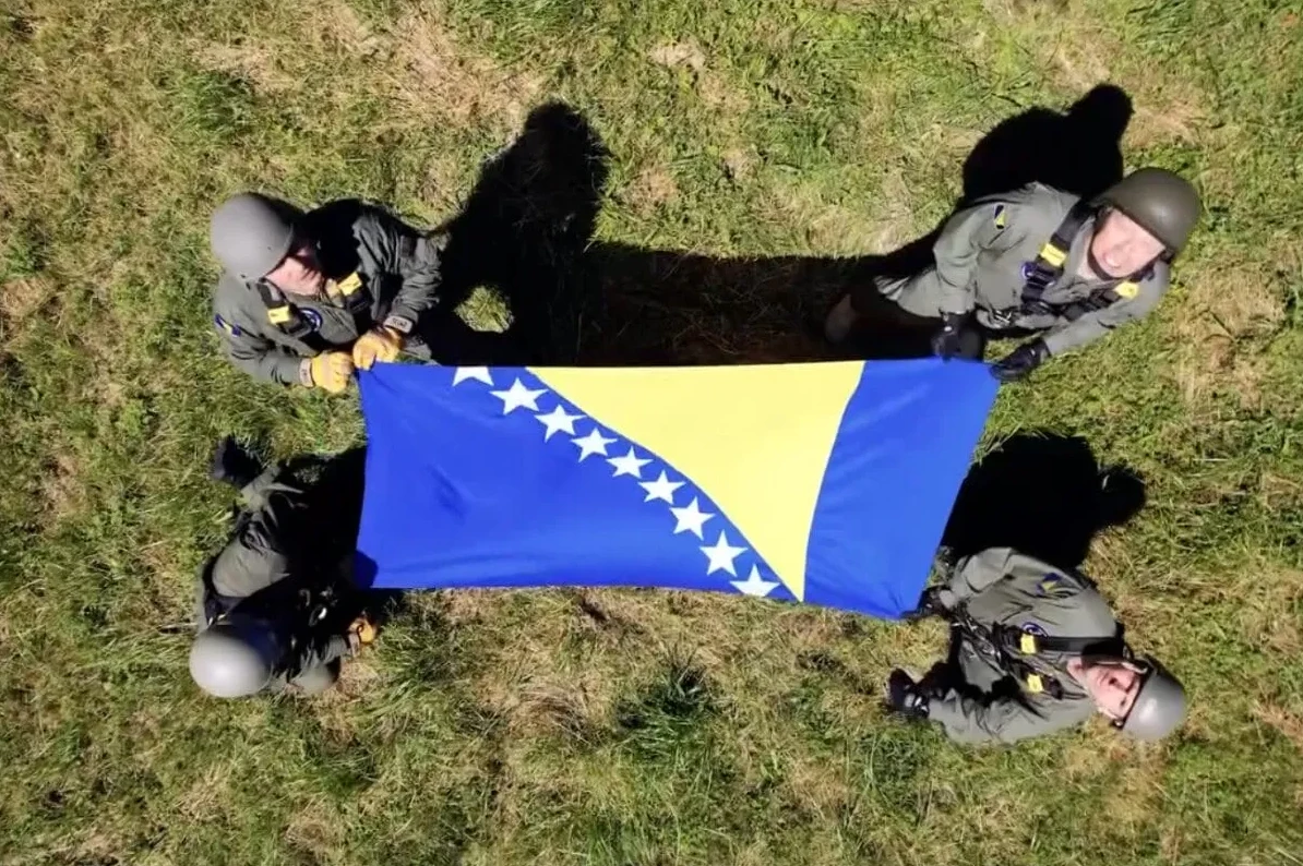 Četiri pilota u uniformama i sa kacigama drže rasklopljenu zastavu Bosne i Hercegovine na travi, gledajući prema njoj.