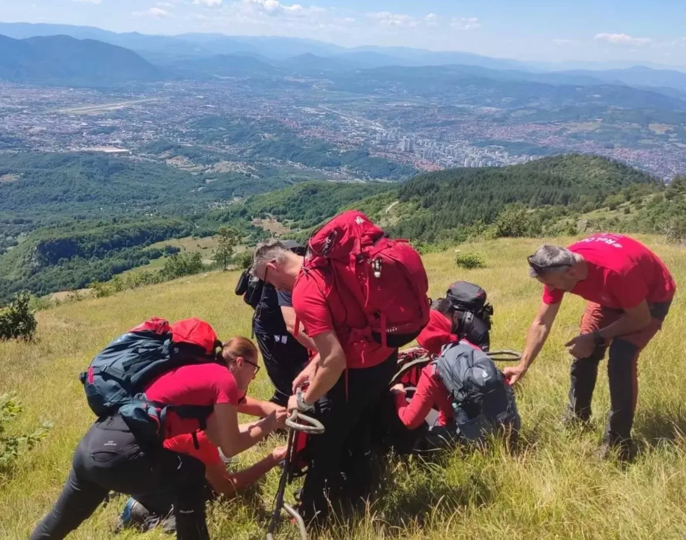 Grupa planinara u crvenim majicama nosi velike rančeve i pregleda opremu na vrhu planine s pogledom na grad u daljini.