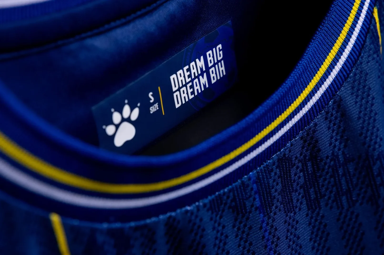 Detaljan prikaz plavog dresa sa natpisom "DREAM BIG DREAM BIH" i logom šape na etiketi veličine "S".