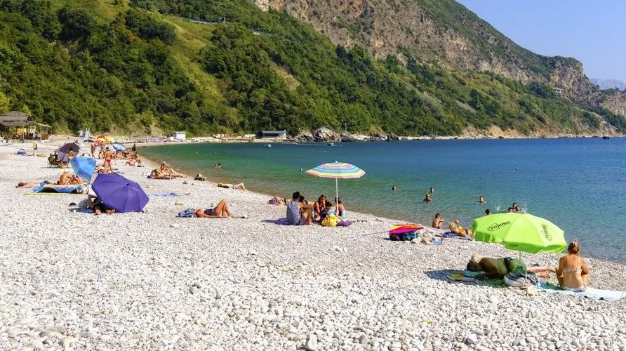Plaža sa šljunkom, ljudi koji se sunčaju i kupaju, suncobrani i planina obrasla zelenilom u pozadini.