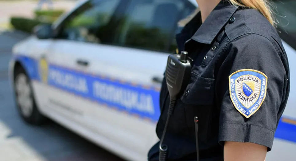 Policajac ili policajka Brčko Distrikta BiH u uniformi sa značkom na rukavu i policijskim radio-uređajem. U pozadini se nazire policijsko vozilo sa natpisom "POLICIJA".