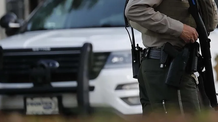 Policajac u uniformi, s pištoljem na pojasu i sačmaricom u ruci, stoji ispred službenog policijskog vozila.