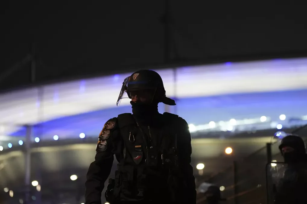 Pripadnik policije u punoj opremi, noseći šljem sa vizirom, stoji u mraku.