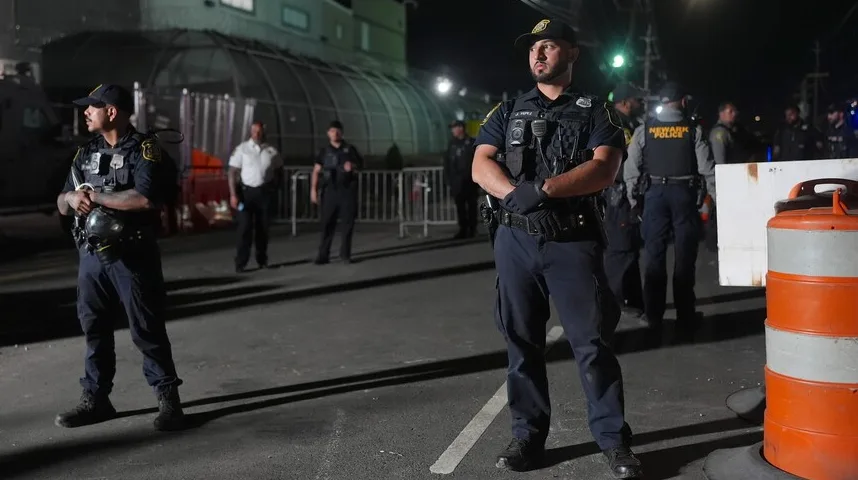 Policajci u uniformi, neki od njih s kacigama u rukama, stoje na ulici noću, osiguravajući neki događaj ili lokaciju.