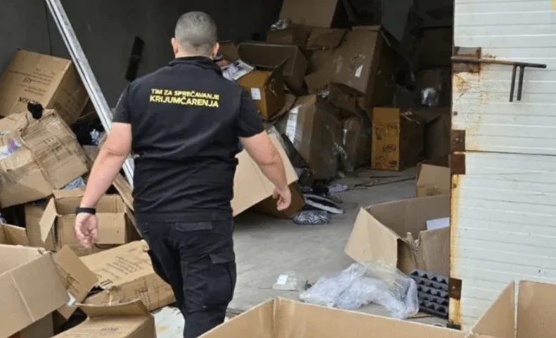 Službenik u crnoj uniformi sa natpisom "Tim za sprečavanje krijumčarenja" pregleda skladište puno kartonskih kutija.