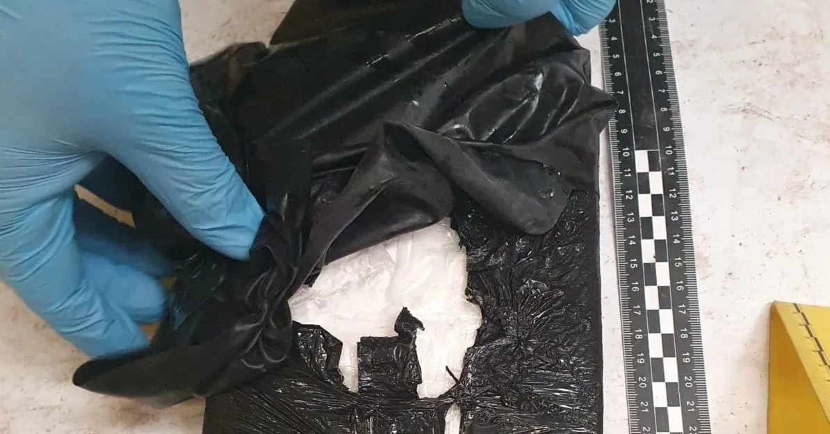 Policajac u plavim rukavicama otvara crnu plastičnu vrećicu u kojoj se vidi bijela materija, pored je postavljeno ravnalo.