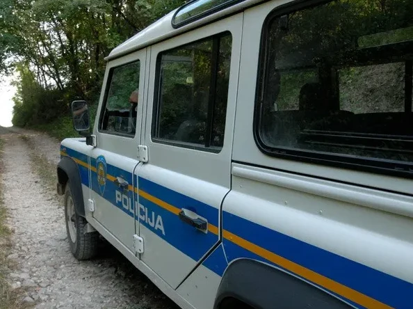 Bijeli policijski džip sa plavim i žutim prugama parkiran na makadamskom putu okružen drvećem.