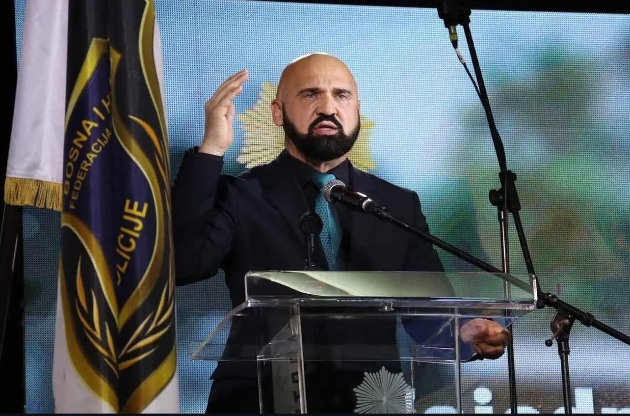 Ćelavi muškarac sa bradom, obučen u tamno odijelo i plavu kravatu, govori iza prozirnog podijuma. Lijevo od njega se nalazi zastava Federacije Bosne i Hercegovine.
