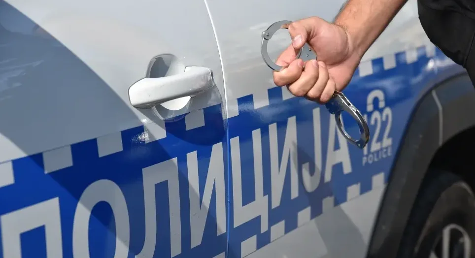 Policajac drži lisice pored službenog policijskog vozila sa natpisom "POLICIJA"