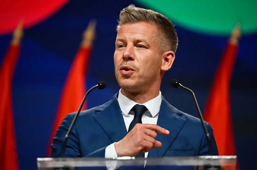 Političar u tamnoplavom odijelu, bijeloj košulji i tamnoj kravati, govori ispred podijuma, s dva mikrofona ispred sebe. U pozadini se naziru tri crvene zastave i plavi panel s crvenim i zelenim elementima.