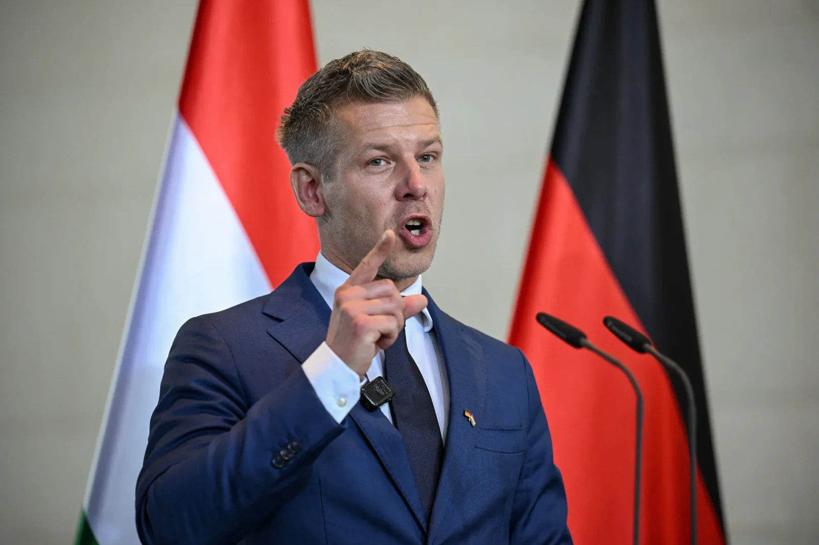 Političar u tamnoplavom odijelu govori, podižući kažiprst dok stoji ispred zastava Njemačke i Mađarske.