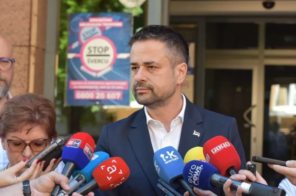Političar u tamnom odijelu, okružen mnoštvom mikrofona medija, drži govor ispred zgrade.