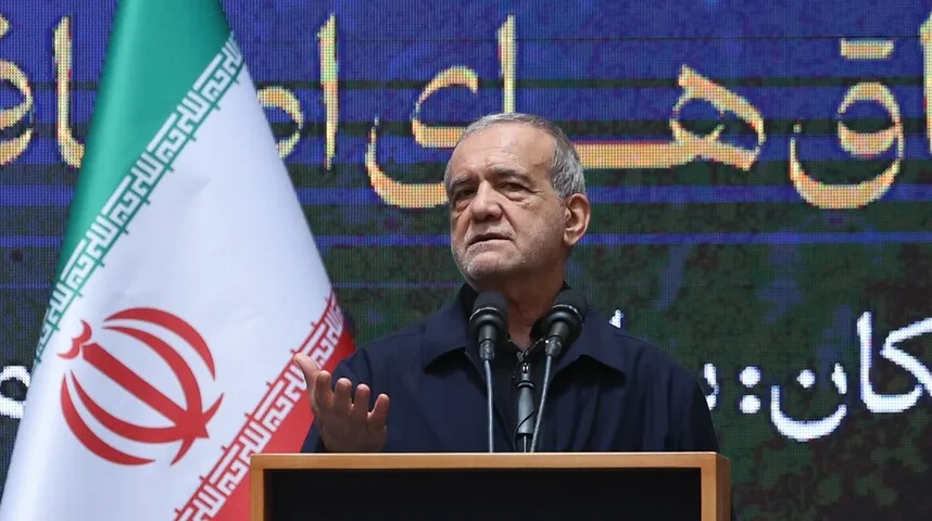 Političar srednjih godina sa sijedom kosom govori sa podijuma, sa iranskom zastavom lijevo i natpisom na perzijskom u pozadini.