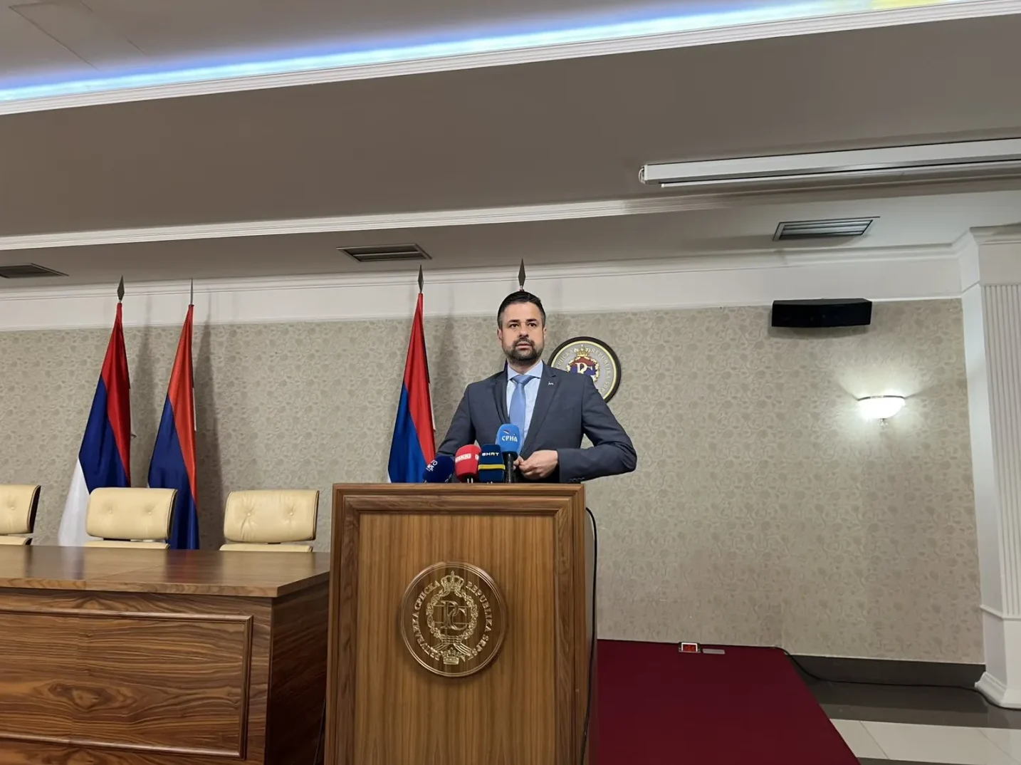 Političar u odijelu stoji iza drvenog podijuma s logom Republike Srpske, a iza njega se nalaze tri zastave i mikrofoni.