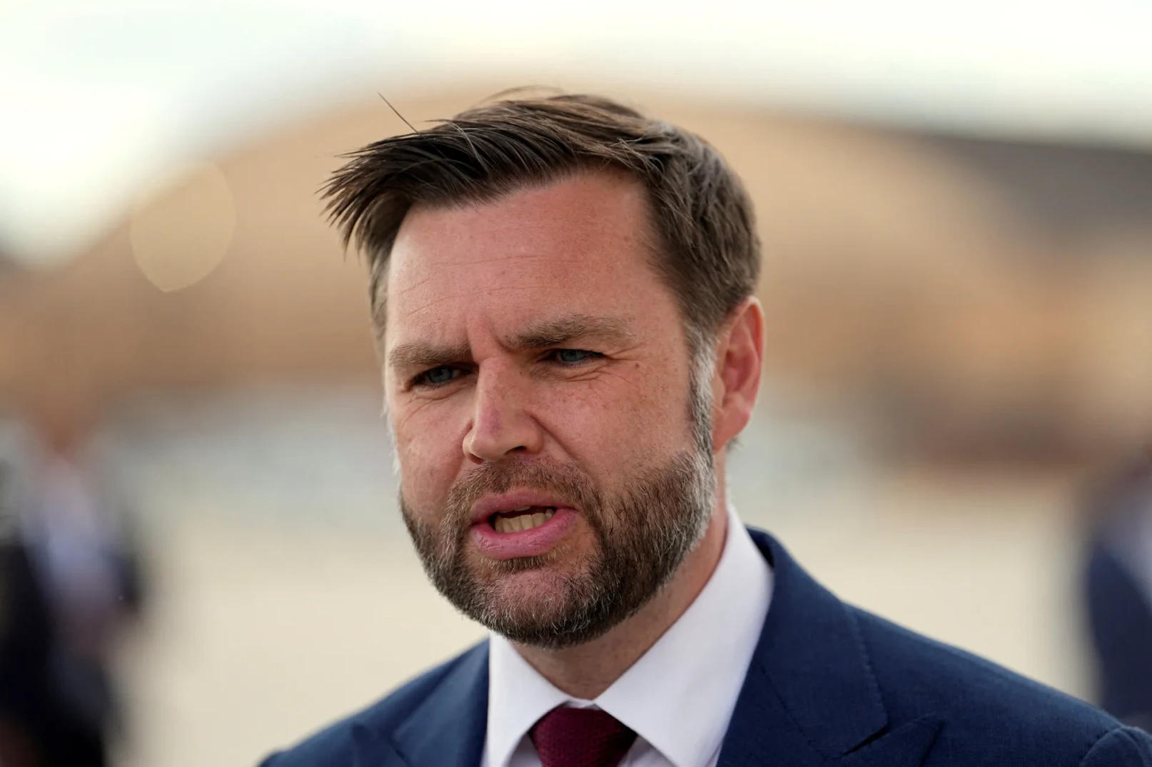 Portret muškarca sa kratkom smeđom kosom, bradom i plavim očima, obučenog u tamno plavo odijelo i bijelu košulju sa crvenom kravatom.