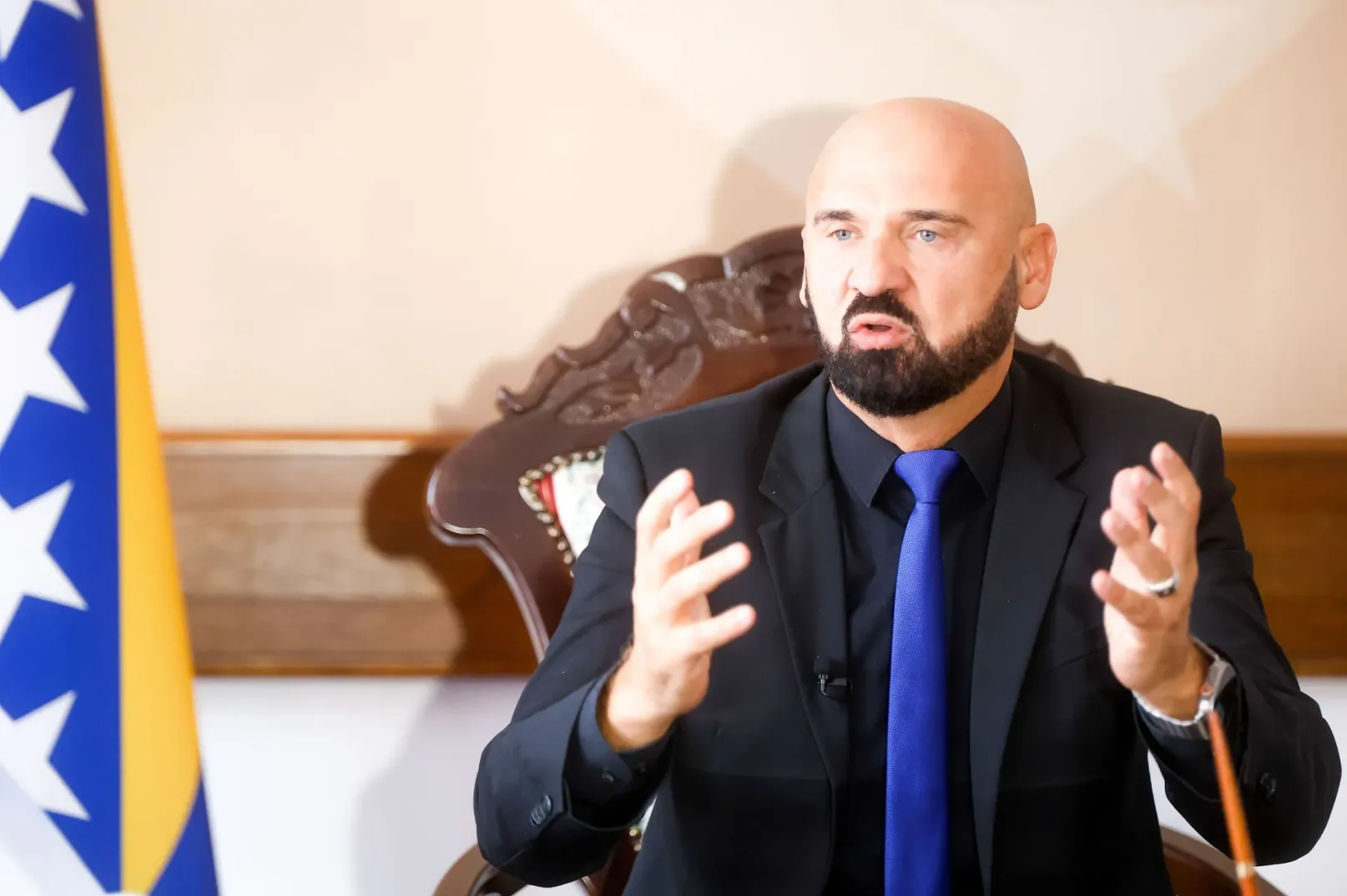 Ćelavi muškarac sa bradom, obučen u crno odijelo i plavu kravatu, sjedi i govori u prostoriji pored zastave Bosne i Hercegovine.